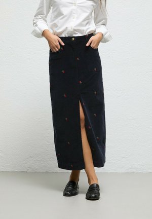 Mujer con una camisa blanca de manga larga, falda midi negra con pequeños detalles florales rojos, abertura frontal y mocasines negros sin cordones.