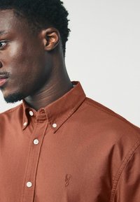 Camicia marrone con bottoni, dotata di colletto, caratterizzata da bottoni bianchi e un logo ricamato sul petto. Tessuto liscio e vestibilità contornata.