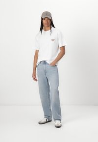 White cotton T-shirt with chest print, light blue denim jeans, and gray cap. Features a relaxed fit and low-top sneakers with black accents.