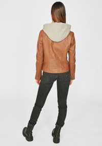 Braune Lederjacke mit grauem Kapuzenaufsatz, taillierte Passform, glatte Textur, kombiniert mit schwarzen Jeans und kniehohen schwarzen Stiefeln.