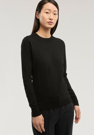 Femme aux cheveux longs et foncés portant un pull à manches longues noir et un pantalon foncé, debout devant un fond gris clair uni.