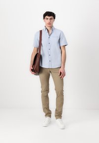 Camicetta azzurra a maniche corte con bottoni e tasca, abbinata a pantaloni beige e sneakers bianche, che tiene in mano una borsa a tracolla in pelle marrone.