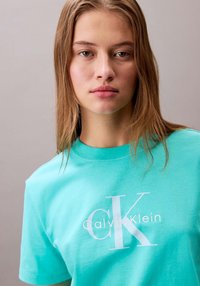 T-shirt di cotone turchese con un grande logo bianco "CK" e la scritta "Calvin Klein". Girocollo e maniche corte.