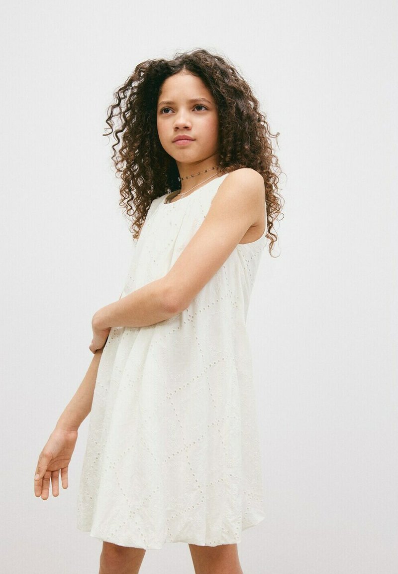 Fille aux cheveux bouclés portant une robe blanche sans manches, debout devant un fond uni, regardant légèrement sur le côté avec une expression neutre.