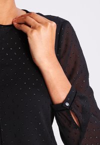 Blusa nera in tessuto trasparente, con perline argentate texturizzate e dettagli con polsini bottonati. Dettaglio ravvicinato del collo e della manica.