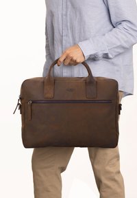 Brun læder arbejdstaske med to top håndtag, en frontlomme med lynlås og en glat overflade. Har subtilt brandlogo prægede på forsiden.