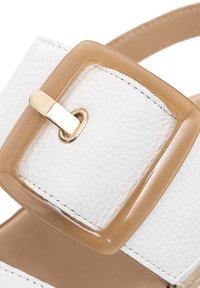 Sandalia de cuero blanco que presenta una gran hebilla transparente color beige con un cierre dorado. Textura suave con detalles de costura en la correa.