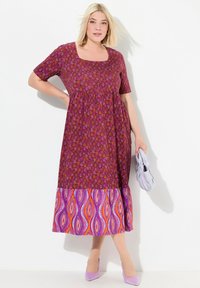 Lila und rosa gemustertes Kleid mit kurzen Ärmeln und quadratischem Ausschnitt, mit einem kontrastierenden gemusterten Saum. Leichte Schuhe und eine graue Tasche sind enthalten.