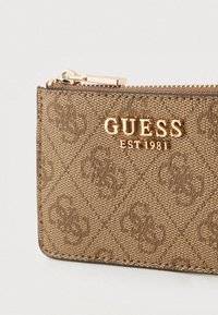 Portafoglio marrone con motivo a fantasia e logo dorato "GUESS EST 1981", superficie testurizzata e chiusura con cerniera. Design compatto adatto per carte e monete.