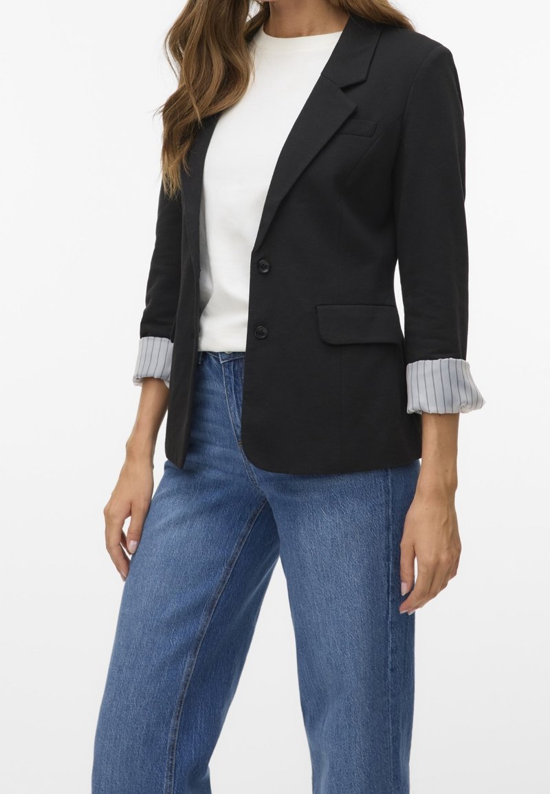 Svart blazer med skräddarsydd design, framknappar och slagkrage. Ärmarna har ett lätt grått randigt tyg. Matchas med blå jeans.