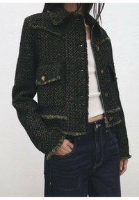 Czarna i zielona kurtka z tweedu o kroju cropped, z postrzępionymi krawędziami, zapinana na guziki, z dwoma przednimi kieszeniami. Noszona na białej bluzce.