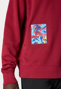 Κόκκινο φούτερ φτιαγμένο από μαλακό ύφασμα με μια ορθογώνια παραλληλεπίπεδη τσέπη που έχει ένα πολύχρωμο αφηρημένο σχέδιο σε μπλε, κόκκινο και κίτρινο.