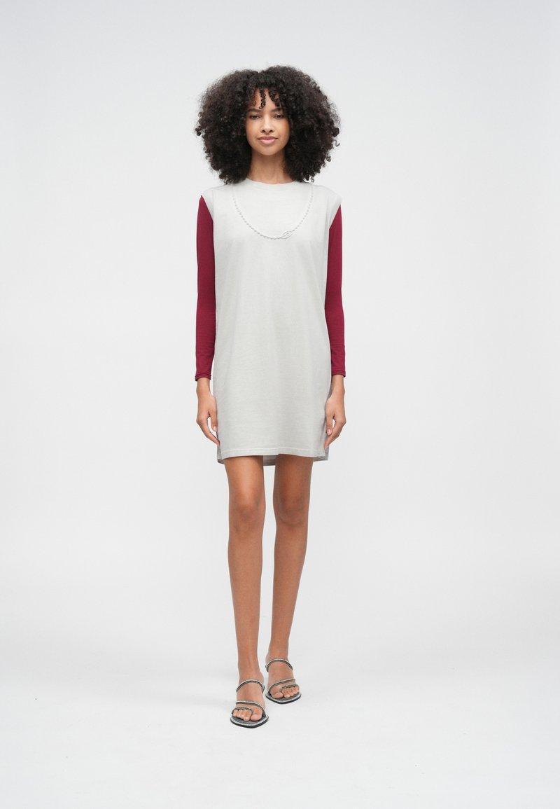 Jauna sieviete ar cirtainiem matiem stāv, valkājot gaiši pelēku kleitu bez piedurknēm pār garām piedurknēm bordo krāsas krekla un ādas sandales ar atvērtām purngalām.