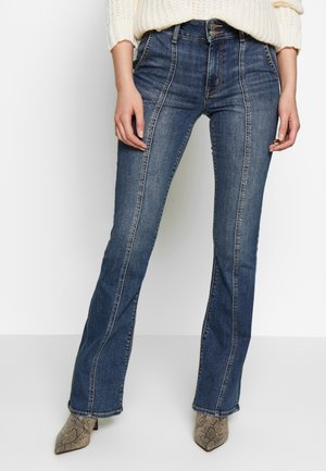 Femme portant un jean en denim bleu taille haute avec coutures avant et jambes évasées, associé à un pull crème et des bottines à imprimé serpent.