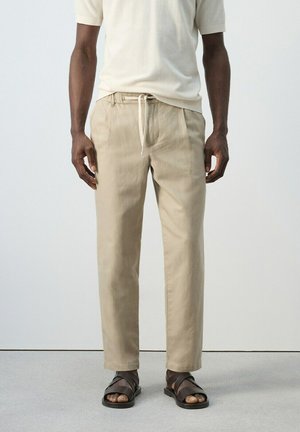 Hombre con pantalones beige con cordón, camiseta de manga corta crema y sandalias marrón oscuro, de pie contra un fondo liso.