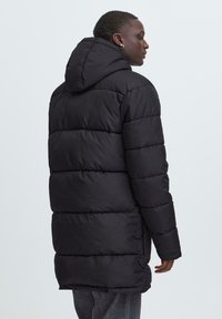 Persona vestida con una larga chaqueta acolchada negra con capucha, girada hacia un lado, sobre un fondo claro y liso.