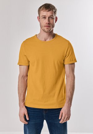 T-shirt giallo senape a maniche corte con scollo rotondo, realizzato in tessuto morbido. Indossato da una persona, abbinato a jeans blu.