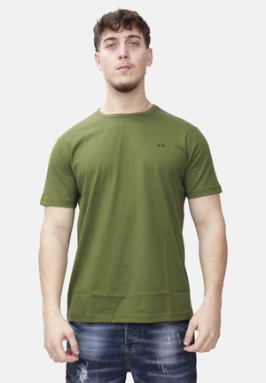 Giovane uomo con capelli corti e ricci che indossa una maglietta a maniche corte verde oliva e jeans blu consumati, in piedi davanti a uno sfondo semplice.