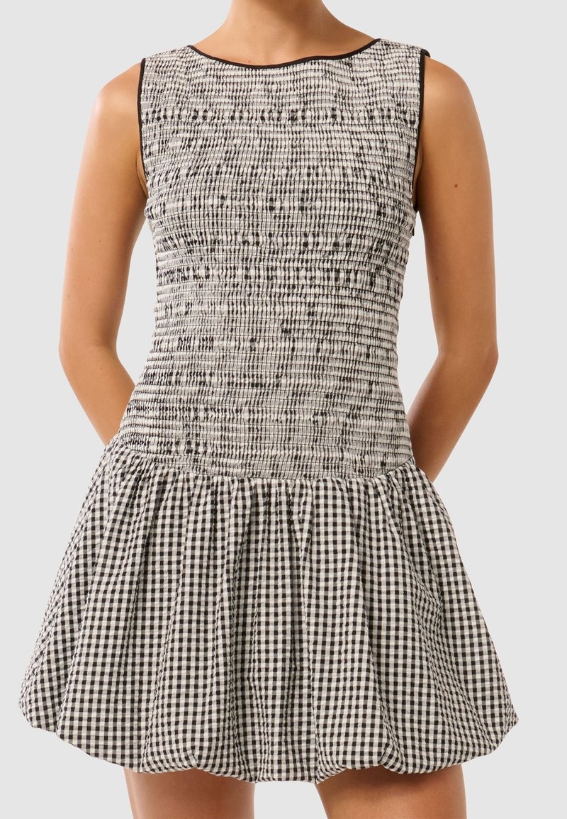 Robe sans manches à carreaux noirs et blancs avec corsage ajusté smocké et jupe évasée portée par une personne debout devant un fond uni.