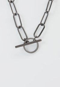 Collier en chaîne métallique avec de grands maillons en finition gris foncé, présentant un pendentif circulaire et un fermoir à barre horizontale.