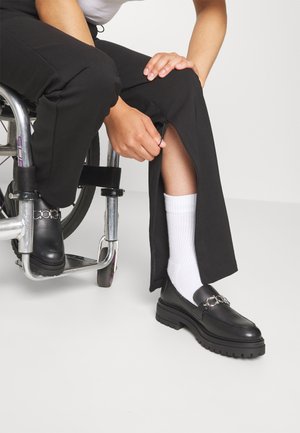 Mocassins en cuir noir avec une semelle épaisse et un détail de chaîne en argent décorative. Associés à un pantalon noir et des chaussettes blanches, assis dans un fauteuil roulant.
