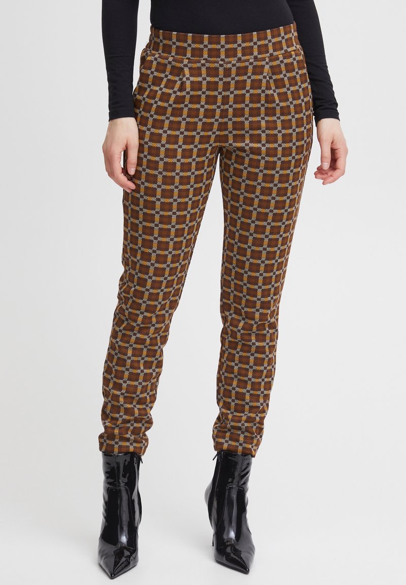 Femme portant un pantalon slim à carreaux marron et beige avec des bottines noires à talons aiguilles pointues, debout sur fond blanc.