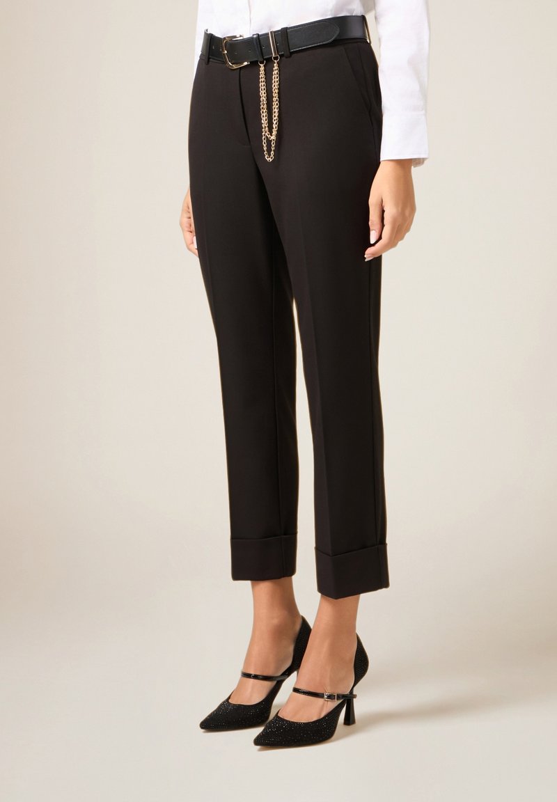 Pantalones negros a medida con dobladillo vuelto, combinados con tacones glitter con correa en el tobillo. Incluyen un cinturón negro con un detalle de cadena dorada.