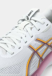 Witte sportschoen met een gemêleerde bovenkant van mesh, oranje en bordeauxrode accenten, platte veters en een roze tussenzool. Merkenlogo op de tong.