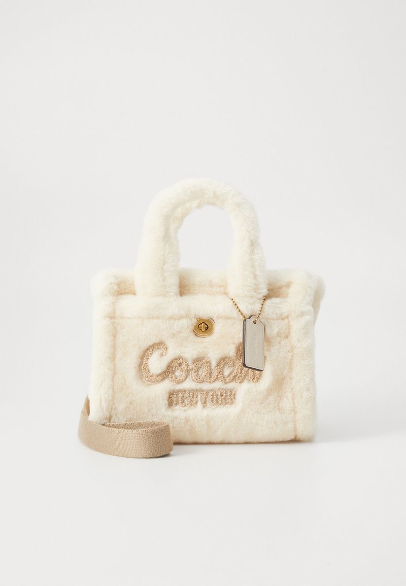 Krém színű plüss Coach kézi táska rövid fogantyúkkal, arany színű záródással, levehető bézs pánttal és függő márkacímkével.