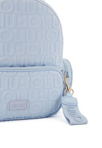 Zaino azzurro chiaro con texture, caratterizzato da un motivo a logo in rilievo, tasca frontale con zip e accenti in metallo dorato. Finitura liscia e materiale resistente.