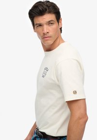 Cremefarvet bomulds-t-shirt med rund halsudskæring, korte ærmer og et blåt trykt logo på brystet. Indeholder et mærke på ærmet.