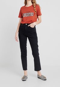 Rust t-shirt med sort og hvid grafisk tekst, parret med sorte straight-leg jeans og stribede slip-on sko.