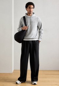 Felpa grigia con coulisse, borsa a tracolla nera, pantaloni neri larghi, sneakers bianche, in piedi su un pavimento di legno contro uno sfondo chiaro.