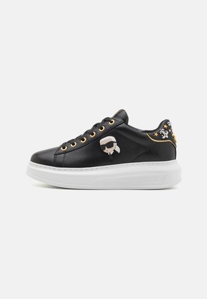 Black leather sneaker with white thick sole, gold eyelets, cartoon figure patch, and silver star and skull embellishments on heel.