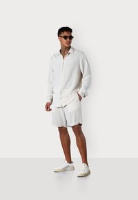 Helle Leinenbluse mit Knopfverschluss und langen Ärmeln, kombiniert mit passenden Shorts. Weiße Sneakers vollenden den Look.