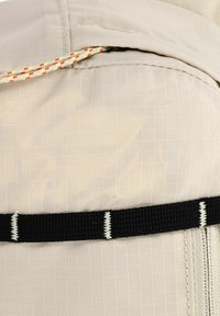 Zaino leggero con un tessuto beige testurizzato, dotato di una tracolla nera e una chiusura con cordino multicolore.