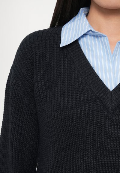 Tengerészkék kötött pulóver V-nyakú kialakítással, texturált felülettel. Világoskék csíkos galléros ing fölé viselve.