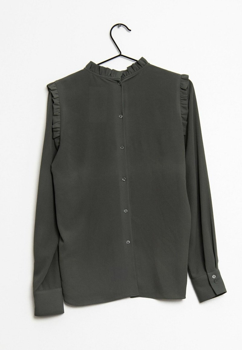 Blouse à manches longues gris foncé avec col à volants et détails aux épaules, boutonnée dans le dos, suspendue à un cintre noir.