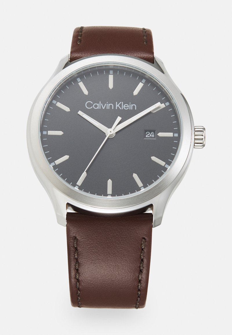 Montre Calvin Klein avec un boîtier en acier inoxydable, un cadran gris foncé, des index en argent et un bracelet en cuir marron avec des coutures visibles.