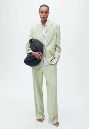 Lichtgroen pak met een op maat gemaakte blazer en broek met wijde pijpen, gecombineerd met een wit overhemd en een gestructureerde zwarte gevlochten tas.