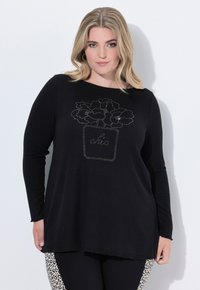 Frau mit blonden Haaren in einem schwarzen Langarmshirt mit floralem Topfdesign und dem Text "chic", kombiniert mit schwarzen und leopardgemusterten Hosen.