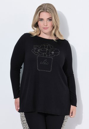 Vrouw met blond haar die een zwarte langwerpige top draagt met een bloemenpot ontwerp en de tekst "chic", gecombineerd met zwarte en panterprint broek.
