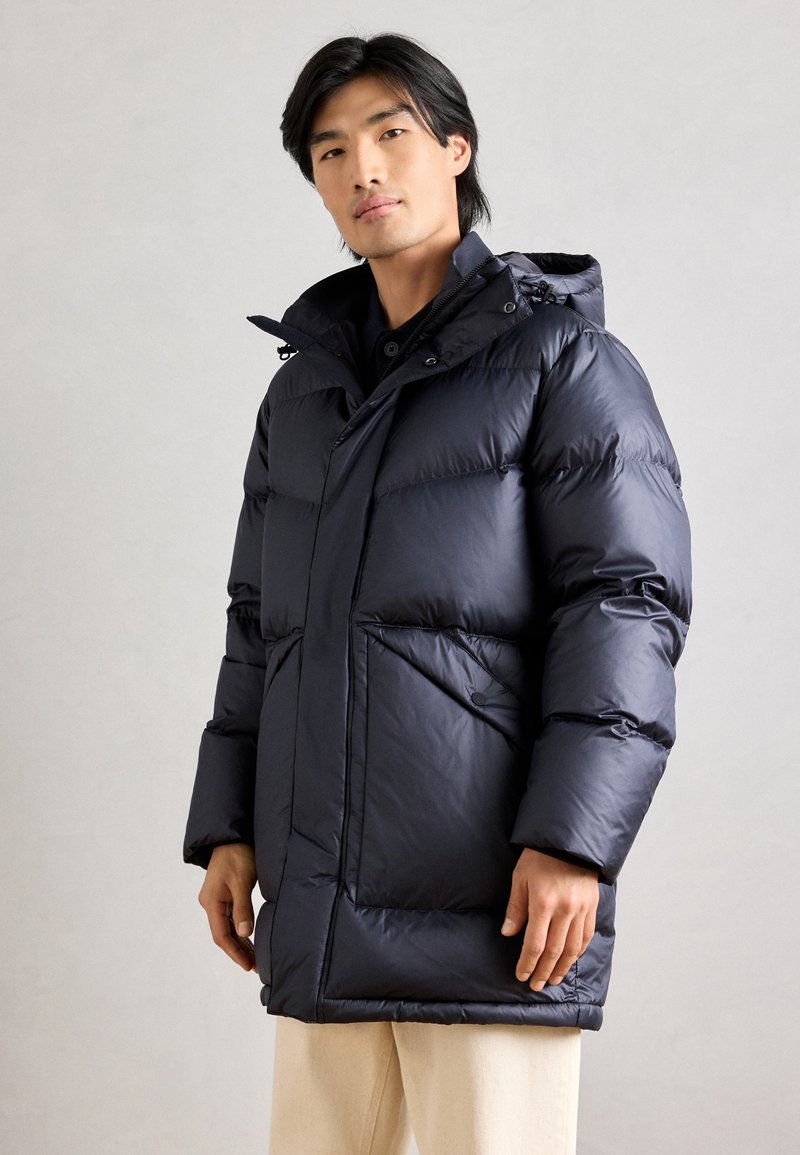 Casaco puffer azul-marinho com colarinho alto, fecho frontal com botões e dois grandes bolsos laterais. Apresenta uma textura suave e brilhante.