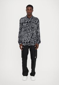 Schwarz-weiß gemustertes Langarmshirt mit Textdesign; getragen mit schwarzen Cargo-Hosen und weißen Sneakers, stehend vor einem schlichten Hintergrund.
