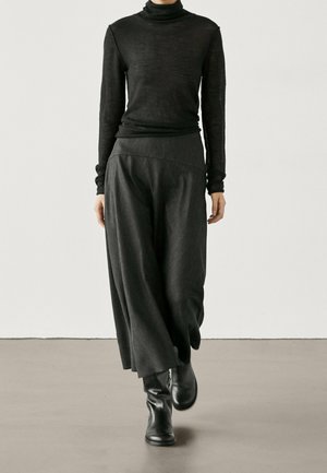 Personne portant un col roulé noir, un pantalon large gris foncé et des bottines noires marchant sur un sol gris contre un mur blanc.