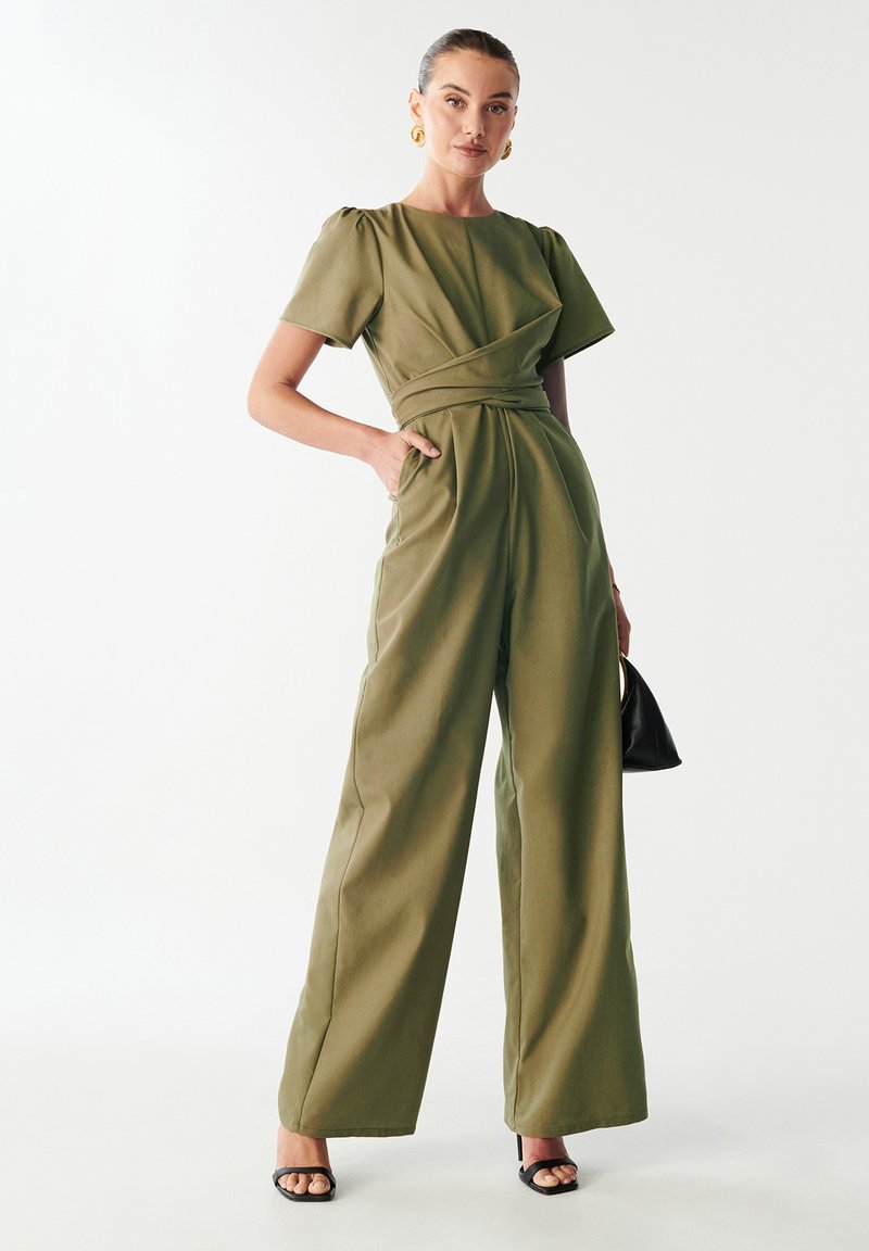 Mono verde oliva con mangas cortas, detalle de cintura envuelta, pantalones de pierna ancha y tejido suave; complementado con un pequeño bolso negro.