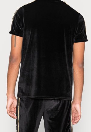 Person iført sort fløjls-top med korte ærmer og matchende bukser med guldfarvede striber i siden, set bagfra med en tatovering på venstre arm.