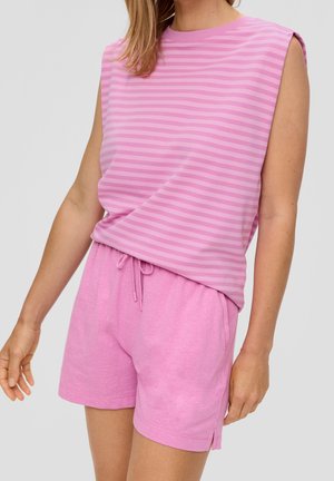 Frau trägt ein rosa gestreiftes ärmelloses Oberteil und rosa Shorts mit Kordelzug, steht mit leicht gebeugtem Arm.