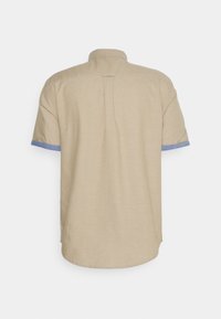 Camisa de manga corta beige hecha de una tela ligera, con detalles de puño de denim y un dobladillo redondeado, con un cuello estándar y un pliegue en la parte posterior.