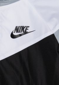 Nike bunda s bílým a černým barevným blokem, s hladkou texturou a výrazným černým logem na hrudi.